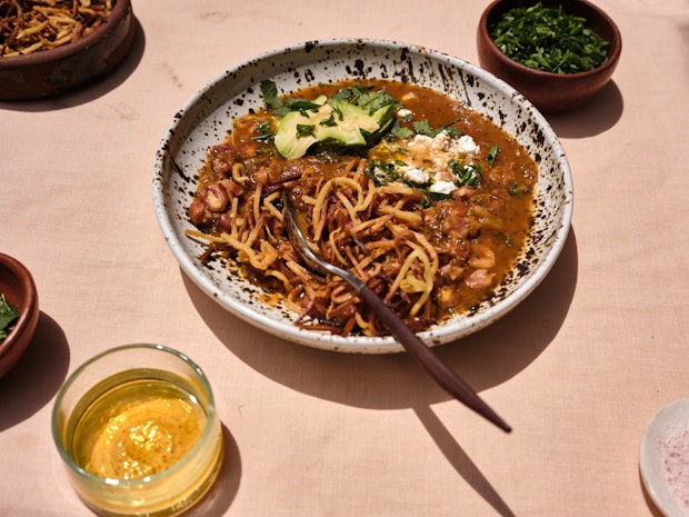 A Close-up of a bowl of Vegan Pozole with toppings including Tortilla Chips, Avocado and Cilantro