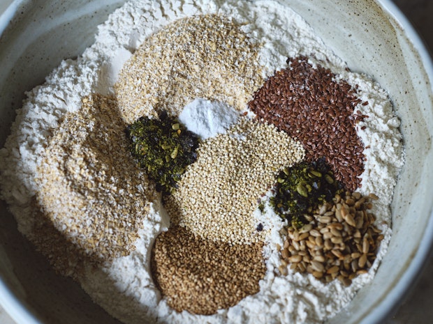 Large mixing bowl with bread ingredients including flax seeds, sunflower seeds, quinoa, flour, and bran