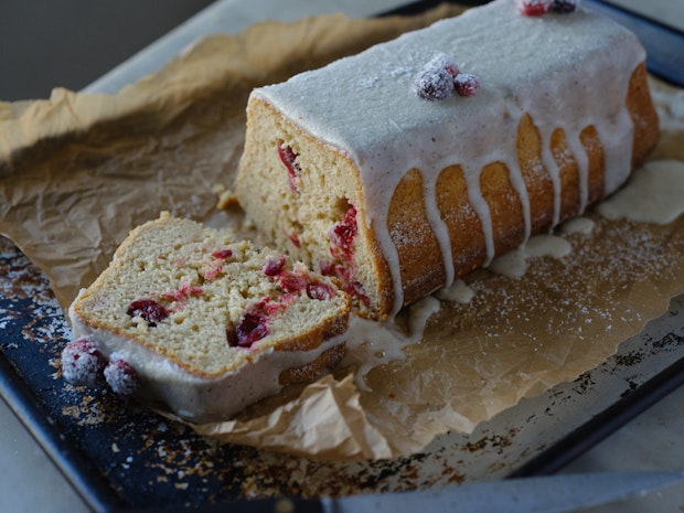 A sliced cranberry cake with vanilla icing with cranberries dotting the crumb of the cake on a sheet of parchment paper