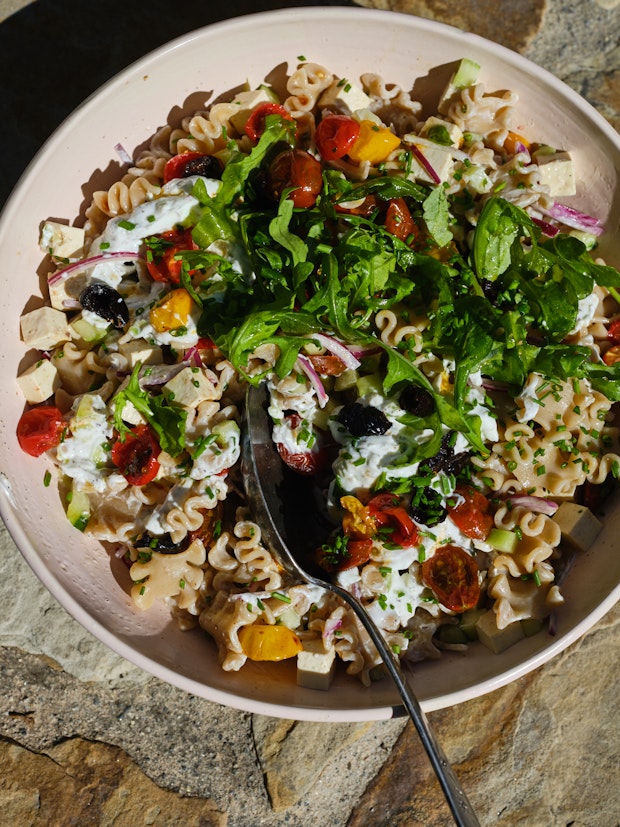 Mediterranean pasta salad with tomatoes, black olives, cucumber, red onions with a creamy yogurt dressing in a wide pink bowl