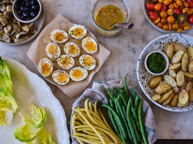 ingredients for a Nicoise arranged on a counter including hard-boiled eggs, green beans, potatoes, olives, lettuces