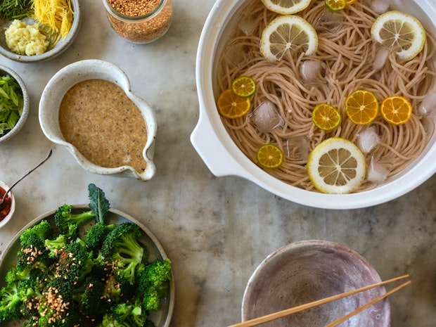 noodles floating in ice water surrounded by toppings and sesame sauce on a table